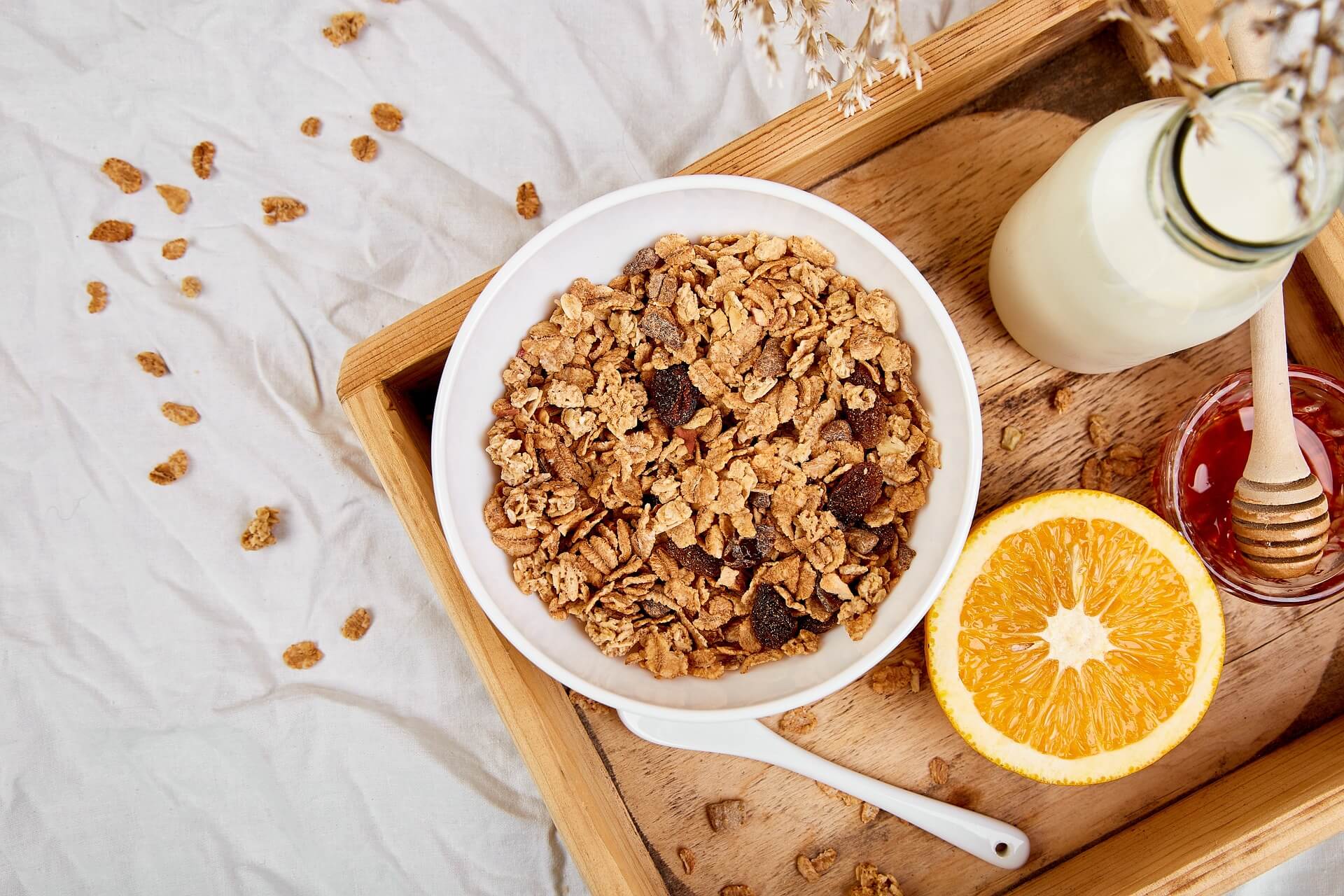 Gedecktes Frühstückstablett mit einer Schüssel frischem Müsli, einer aufgeschnittenen Orange, einer Glasflasche Milch und einem Honigtopf mit Honiglöffel.