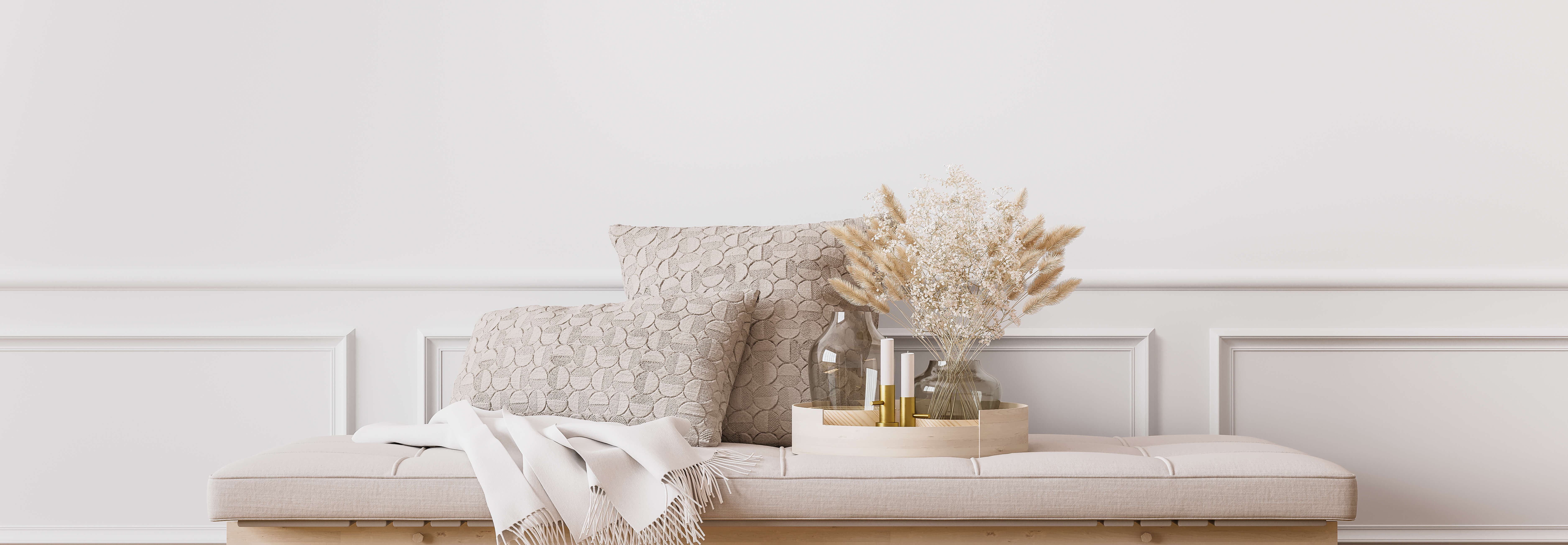 Dekorative Kissen, eine Decke und eine Glasvase mit getrockneten Gräsern drapiert auf einer weiß gekalkten Oberfläche vor einer weißen Paneelwand.