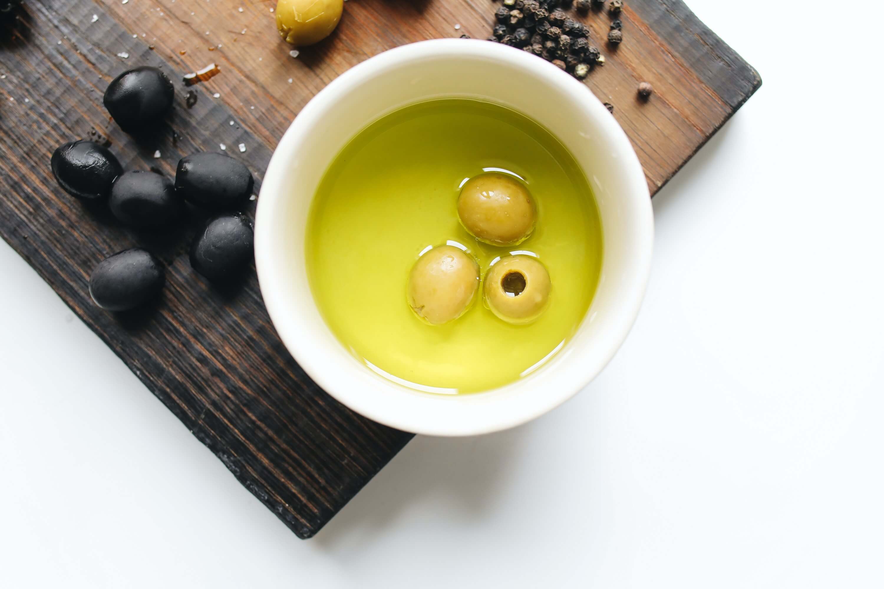Weiße Schüssel mit Olivenöl und grünen Oliven auf einem Holzbrett, ergänzend zum Blogbeitrag 'Der ultimative Guide zu Olivenöl' auf Regizon